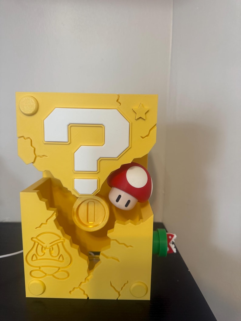 Pode incluir: Um bloco amarelo com um ponto de interroga&ccedil;&atilde;o branco, um cogumelo vermelho e branco e uma moeda de ouro. O bloco tem um design Goomba e uma estrela. O objeto &eacute; impresso em 3D.