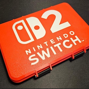 May include: A bright red Nintendo Switch case with the Nintendo Switch logo and the number 2 in white. The case has black latches and a textured surface.