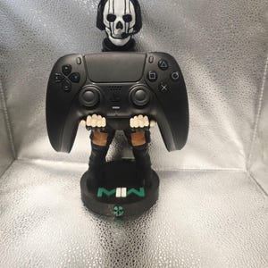 K&ouml;nnte beinhalten: Ein schwarzer PlayStation 5 Controller-St&auml;nder mit einem sch&auml;delgesichtigen Charakter aus dem Videospiel Call of Duty: Modern Warfare II. Der St&auml;nder hat eine schwarze Basis mit den Buchstaben "MWII" in Gr&uuml;n. Der Controller wird von den Z&auml;hnen des Charakters gehalten.