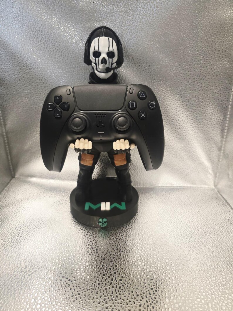 K&ouml;nnte beinhalten: Ein schwarzer PlayStation 5 Controller-St&auml;nder mit einem sch&auml;delgesichtigen Charakter aus dem Videospiel Call of Duty: Modern Warfare II. Der St&auml;nder hat eine schwarze Basis mit den Buchstaben "MWII" in Gr&uuml;n. Der Controller wird von den Z&auml;hnen des Charakters gehalten.