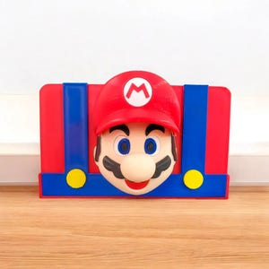 May include: A red, blue, and yellow Mario-themed decorative item. The item features a red hat with a white 'M' and a smiling Mario face. The background is red with blue suspenders and yellow buttons. The item is placed on a wooden surface.