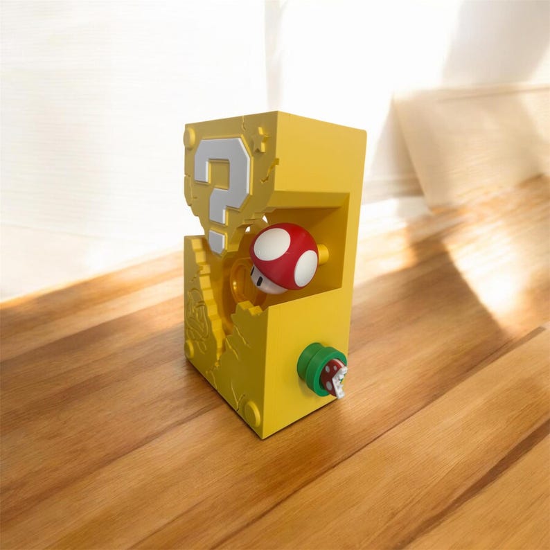 Pode incluir: Uma caixa amarela retangular com um ponto de interroga&ccedil;&atilde;o branco e um cogumelo vermelho e branco. Uma planta verde e vermelha se estende da lateral. O objeto &eacute; um item de novidade, provavelmente um brinquedo ou colecion&aacute;vel.