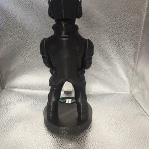 K&ouml;nnte beinhalten: Eine schwarze Figur, die eine Person in taktischer Ausr&uuml;stung darstellt, einschlie&szlig;lich Helm und K&ouml;rperpanzer. Die Figur steht auf einem kreisf&ouml;rmigen Sockel mit einem gr&uuml;n-wei&szlig;en Streifen. Das Design ist detailliert und scheint 3D-gedruckt zu sein.