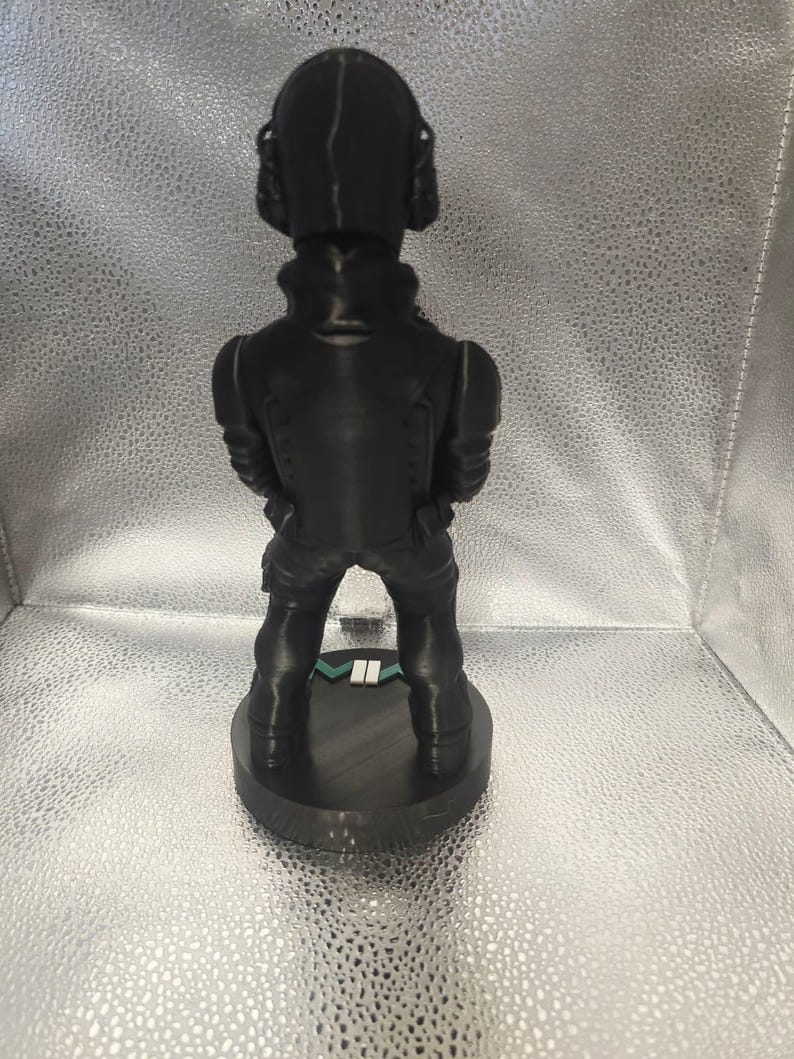 K&ouml;nnte beinhalten: Eine schwarze Figur, die eine Person in taktischer Ausr&uuml;stung darstellt, einschlie&szlig;lich Helm und K&ouml;rperpanzer. Die Figur steht auf einem kreisf&ouml;rmigen Sockel mit einem gr&uuml;n-wei&szlig;en Streifen. Das Design ist detailliert und scheint 3D-gedruckt zu sein.