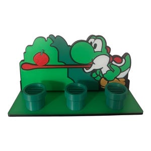 May include: A green and white Yoshi character figure with a red apple on a green background. Three green pipes are in front of the character. The figure is on a green base.