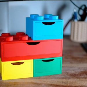 Puede incluir: Una pila de cuatro cajones de almacenamiento coloridos diseñados para parecerse a grandes bloques de juguete. Los cajones son de colores primarios: rojo, azul, amarillo y verde. Cada cajón tiene un pequeño tirador curvo.