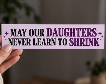 May include: A rectangular light purple sticker featuring the black and dark purple text "MAY OUR DAUGHTERS NEVER LEARN TO SHRINK" in a bold serif font, accented by small four-pointed star icons on the far left and right sides of the message.