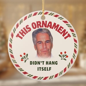 Może przedstawiać: Okrągła ceramiczna ozdoba świąteczna z portretem i napisem "THIS ORNAMENT DIDN'T HANG ITSELF". Ozdoba ma czerwono-zieloną obwódkę w paski i złotą sznurkę do zawieszenia. Ozdobne gałązki znajdują się na dole.