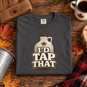 May include: A charcoal gray short-sleeve t-shirt featuring a graphic of a maple syrup jug with a maple leaf icon and the text I'D TAP THAT in bold white and brown lettering. The shirt has a Comfort Colors brand label at the inner neckline.