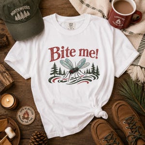 May include: A white short-sleeve t-shirt featuring a graphic of a mosquito above a stylized forest and water scene. The text "Bite me!" is printed in red above the insect. The shirt has a crew neckline and a label at the inner collar area.