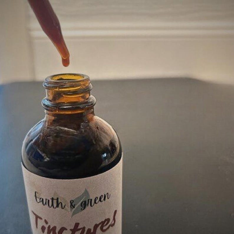 May include: A brown glass tincture bottle with a dropper dispensing a dark liquid. The bottle has a white label with the words "Earth & green Tinctures" in a script font. The bottle is partially filled with a dark liquid.