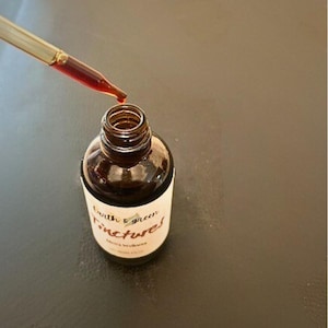 May include: A brown glass bottle labelled "Earth & Green Tinctures" is being filled with a red liquid from a dropper. The bottle sits on a dark surface. The image focuses on the bottle and dropper, highlighting the product.