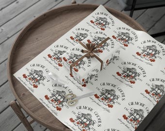 May include: A wrapped gift with a brown ribbon sits on a sheet of patterned wrapping paper. The paper and gift box feature a repeating design with the text "GILMORE GHOULS" and a spooky house illustration. A pair of gold scissors is also visible.
