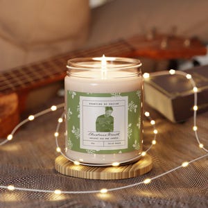 May include: A green and white candle with a label that reads "Sparklins by Design" and "Christmas Wreath" with a picture of a person wearing a hat and scarf. The candle is on a wooden coaster and surrounded by white fairy lights.