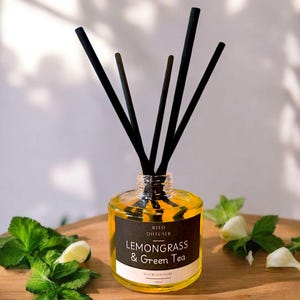 May include: A glass reed diffuser with black reeds, filled with a yellow liquid. The label reads "Lemongrass & Green Tea." Fresh green leaves and a lime wedge are arranged around the diffuser on a wooden surface.