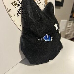 Op de afbeelding: Zwart vilt kattenhoofd met blauwe ogen en witte sterren geborduurd op het gezicht.