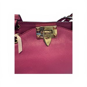 May include: Close-up of a magenta leather handbag. The bag features a gold-colored clasp with pyramid-shaped studs. A white tag with a ribbon is attached to the bag. The bag's design includes a flap and a top handle.