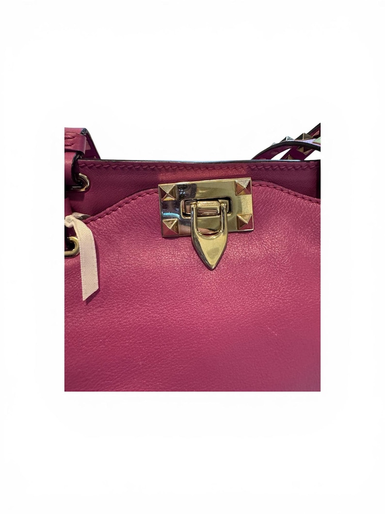 May include: Close-up of a magenta leather handbag. The bag features a gold-colored clasp with pyramid-shaped studs. A white tag with a ribbon is attached to the bag. The bag's design includes a flap and a top handle.
