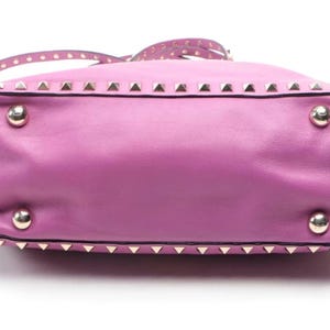 May include: A lavender leather handbag with silver-tone pyramid studs along the top and bottom edges. The bag has a matching strap with small, round studs. Four silver-tone metal feet are visible on the bottom.