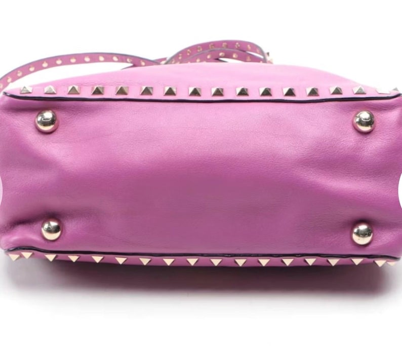 May include: A lavender leather handbag with silver-tone pyramid studs along the top and bottom edges. The bag has a matching strap with small, round studs. Four silver-tone metal feet are visible on the bottom.