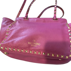 May include: A pink leather tote bag with gold-colored stud accents along the edges and straps. The bag features the "Valentino Garavani" logo in gold lettering. The handles are also studded.