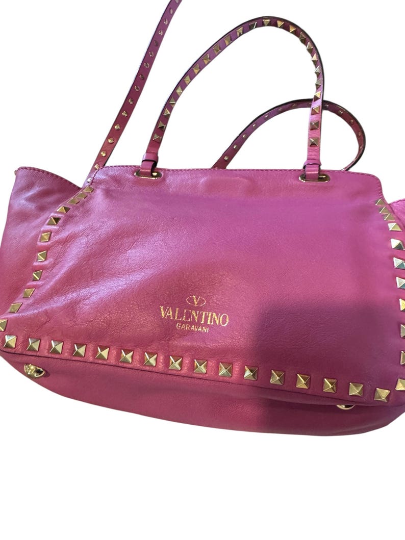 May include: A pink leather tote bag with gold-colored stud accents along the edges and straps. The bag features the "Valentino Garavani" logo in gold lettering. The handles are also studded.