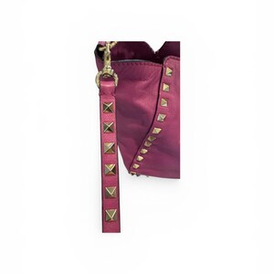 May include: A magenta leather handbag with gold-colored pyramid studs and a matching strap. The strap is adorned with gold-colored studs. The bag has a scalloped top edge and a gold-colored clasp.