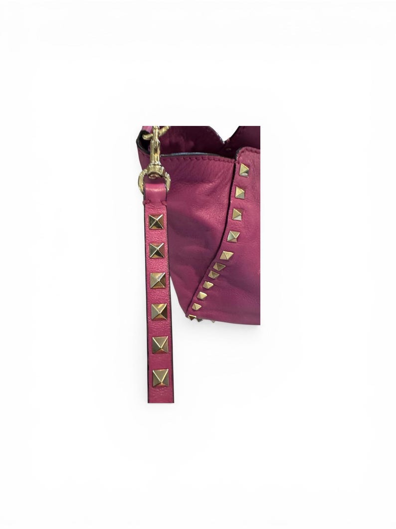 May include: A magenta leather handbag with gold-colored pyramid studs and a matching strap. The strap is adorned with gold-colored studs. The bag has a scalloped top edge and a gold-colored clasp.