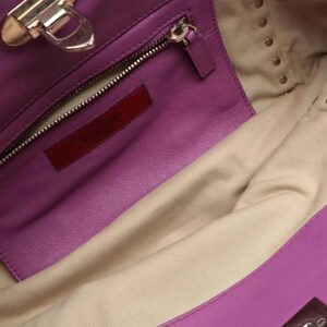 May include: Close-up of a purple leather handbag interior. The bag features a zippered pocket, a red leather tag with "VALENTINO" embossed, and gold-toned hardware. The interior lining is a light beige fabric.