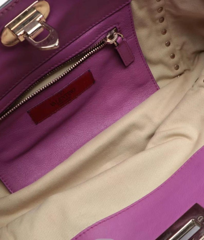 May include: Close-up of a purple leather handbag interior. The bag features a zippered pocket, a red leather tag with "VALENTINO" embossed, and gold-toned hardware. The interior lining is a light beige fabric.