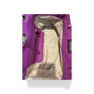 May include: Open view of a bright purple leather handbag. The interior is a light beige color with a zippered pocket and studded accents. Silver-toned hardware is visible on the bag's exterior.