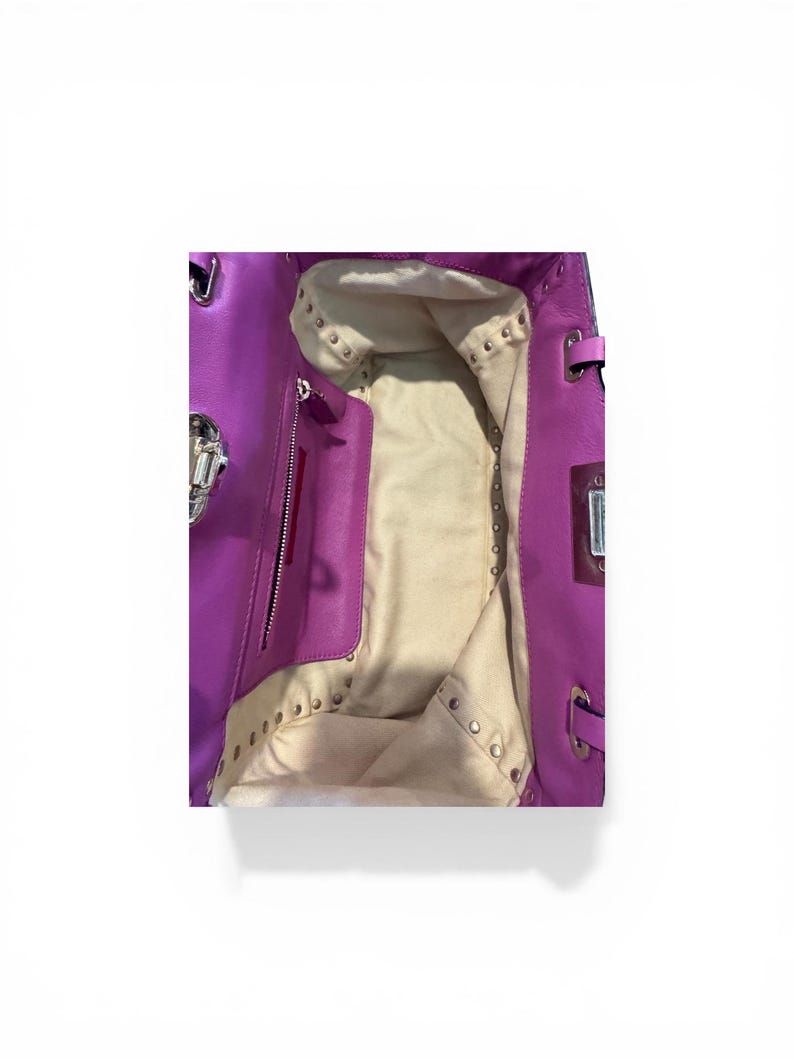 May include: Open view of a bright purple leather handbag. The interior is a light beige color with a zippered pocket and studded accents. Silver-toned hardware is visible on the bag's exterior.