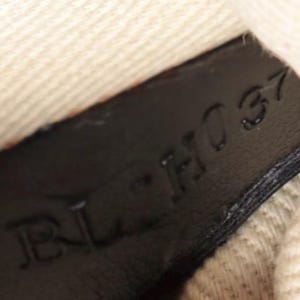 May include: Close-up of a black leather tag with the embossed text "BL-H037". The tag is set against a textured, off-white fabric and a purple zipper.