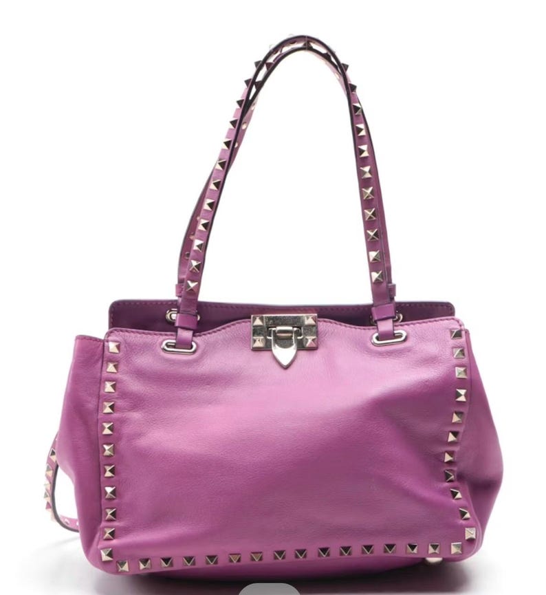 May include: A vibrant purple leather handbag with silver-tone pyramid studs along the edges and straps. The bag features a top handle design, a flap closure, and a structured silhouette, perfect for carrying essentials.