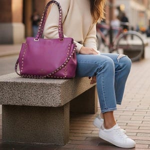 May include: A deep purple handbag with studded accents and a shoulder strap sits on a stone bench. The bag is paired with a cream-colored sweater, blue jeans, and white sneakers. The background shows a city street.