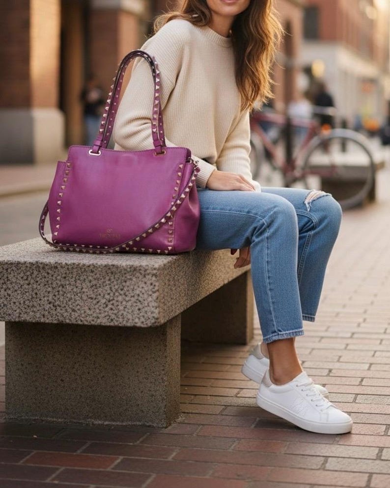 May include: A deep purple handbag with studded accents and a shoulder strap sits on a stone bench. The bag is paired with a cream-colored sweater, blue jeans, and white sneakers. The background shows a city street.