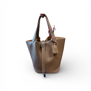 Puede incluir: Bolso tipo cubo de cuero color beige con asa superior y bandolera extraíble. El bolso presenta un cierre de hebilla y una pequeña etiqueta de cuero. El bolso está sostenido por una mano sobre un fondo blanco.