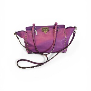 May include: A two-tone purple handbag with gold-colored studs and hardware. The bag has a top handle and a detachable shoulder strap. The bag is made of a leather-like material and has a rectangular shape.