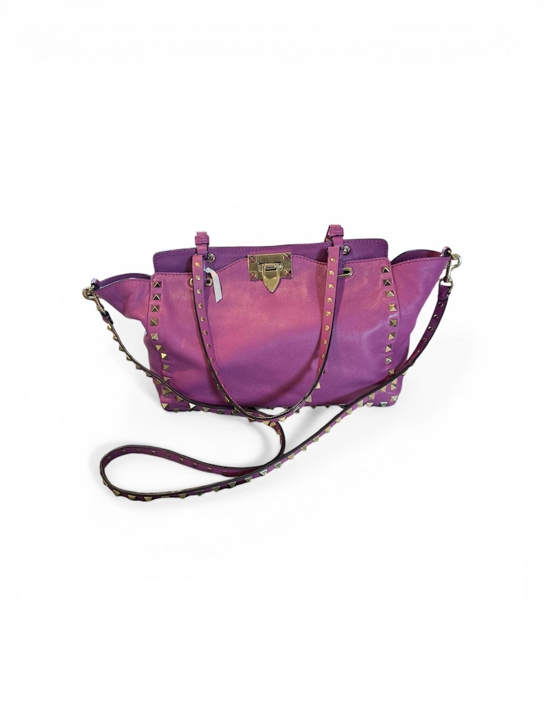 May include: A two-tone purple handbag with gold-colored studs and hardware. The bag has a top handle and a detachable shoulder strap. The bag is made of a leather-like material and has a rectangular shape.
