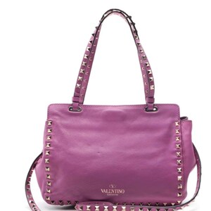 May include: A vibrant purple leather handbag with studded accents. The bag features a top handle and a detachable shoulder strap, both adorned with silver-toned studs. The Valentino Garavani logo is visible on the front of the bag.