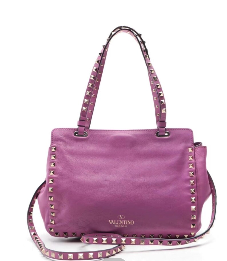 May include: A vibrant purple leather handbag with studded accents. The bag features a top handle and a detachable shoulder strap, both adorned with silver-toned studs. The Valentino Garavani logo is visible on the front of the bag.