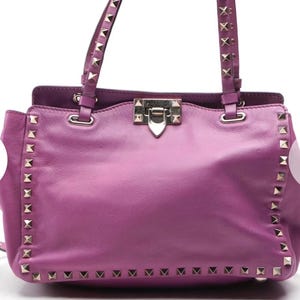 May include: A medium-sized, purple leather handbag with silver-colored stud accents. The bag features two top handles with studded detailing and a silver clasp closure. The bag's exterior is a solid purple color.