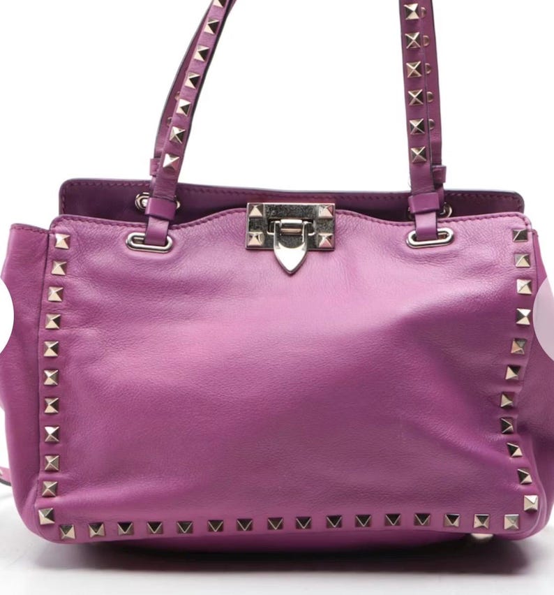 May include: A medium-sized, purple leather handbag with silver-colored stud accents. The bag features two top handles with studded detailing and a silver clasp closure. The bag's exterior is a solid purple color.