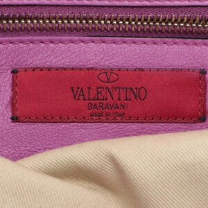 May include: Close-up of a purple leather handbag with a zipper and a red rectangular label. The label features the brand name "VALENTINO GARAVANI MADE IN ITALY" in white lettering. The bag's interior is visible, showing a light-colored fabric.