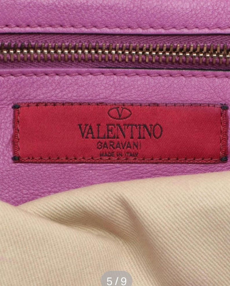 May include: Close-up of a purple leather handbag with a zipper and a red rectangular label. The label features the brand name "VALENTINO GARAVANI MADE IN ITALY" in white lettering. The bag's interior is visible, showing a light-colored fabric.