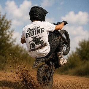 Puede incluir: Una camiseta blanca con el texto "Rather be RIDING" sobre un gráfico de una moto de cross. La camiseta la lleva un piloto con casco negro, pantalones negros y botas blancas, realizando un caballito en una moto negra en una pista.
