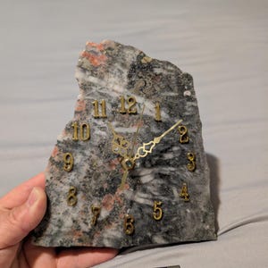 May include: A unique clock made from a slab of marbled stone. The clock face features gold-colored numerals and hands. The stone has a mix of gray, white, and pink hues. A black lighter with the word "especial" is in the foreground.