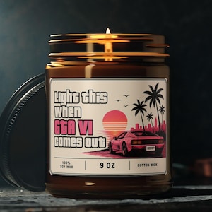 May include: A brown glass jar candle with a lit wick and a label featuring a pink sports car, palm trees, and a sunset. The text reads: Light this when GTA VI comes out. The label also specifies 100% soy wax, 9 oz, and cotton wick.