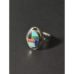 May include: Silver ring with an oval face, showcasing a mosaic design. The mosaic incorporates turquoise, black, red, green, and blue stones. The ring has a detailed band, with a decorative pattern.