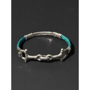 May include: A silver bracelet featuring a central horse bit design connected to two teal-colored leather segments. The metal components are finished with a matte texture, and the inner silver bar is clearly stamped with the text "MEXICO 925".